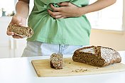 Wenn bei Zöliakie Gluten nicht die einzige Ursache für Verdauungsbeschwerden ist, sollte auch an die Bauchspeicheldrüse gedacht werden. Hier ist ein Mensch zu sehen, der offensichtlich Bauchschmerzen hat. Er hält ein Stück Vollkornbrot in der Hand. Auf dem Tisch vor ihm ist der Rest des Vollkornbrots zu sehen. Bei Menschen mit Zöliakie kann Gluten nicht die einzige Ursache für Verdauungsbeschwerden sein. Es sollte auch an die Bauchspeicheldrüse gedacht werden, da eine begleitende exokrine Pankreasinsuffizienz bei Menschen mit Zöliakie möglich sein kann.